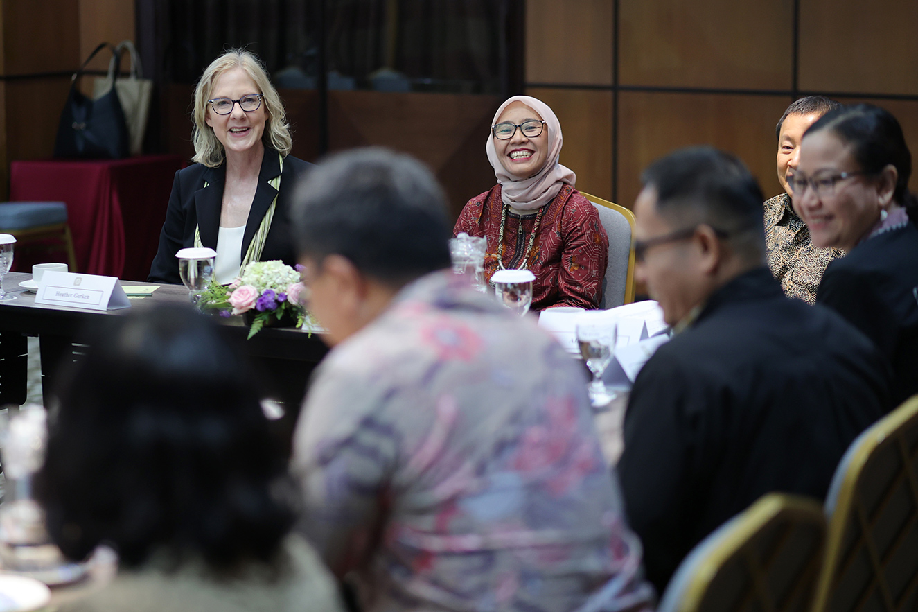 A high-quality photo of professional staff members seated around a conference table in Jakarta, listening to a woman in glasses and a black blazer speak. This action-oriented, journalistic photo captures an engaging meeting that humanizes our global team.