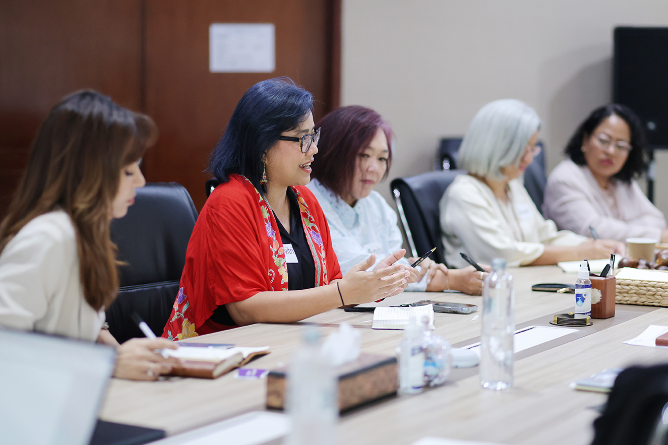 A woman with glasses and a red patterned wrap gestures while speaking at a long conference table. Other professional women listen and take notes during a meeting. This high-quality photo humanizes our global team and highlights collaborative dialogue in a regional setting.