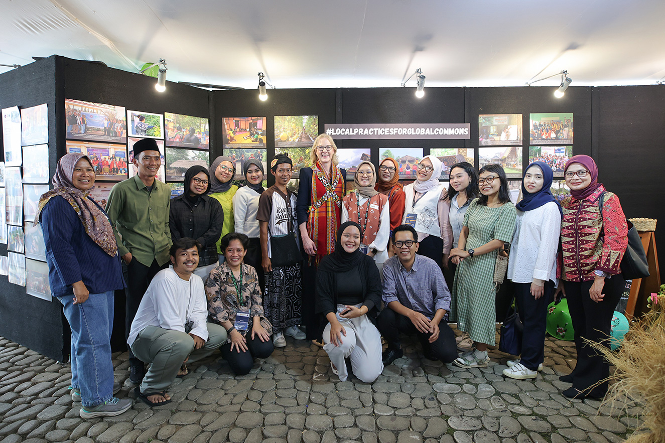 A large, diverse group of over 15 people stands and poses together in front of a black exhibition wall displaying various photographs under the hashtag #LOCALPRACTICESFORGLOBALCOMMONS. This high-quality photo humanizes the foundation's collaborative community engagement in Jakarta, Indonesia.