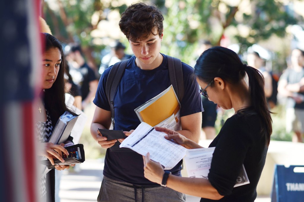 An Advancement Project volunteer reviews voter registration paperwork or community documents with two young college students outdoors on a campus, highlighting legal strategy and grassroots community power in action.