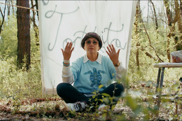 Woman in glasses, a blue t-shirt with a turtle graphic, white button up, and a hat raising her hands while sitting cross-legged outdoors in front of a white banner with text on it.