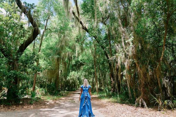 A Black woman in a long and flowing, blue dress stands on a dirt pathway surrounded by a forest of tall, mossy trees. Her beaded hair drapes over her eyes, concealing most of her face.