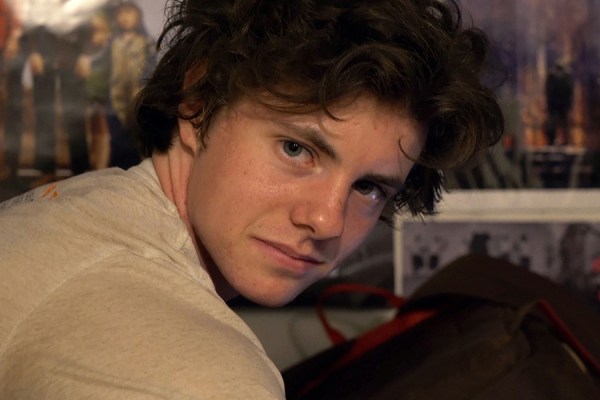 Close-up portrait of a young man with dark short wavy hair looking over his shoulder toward the camera.