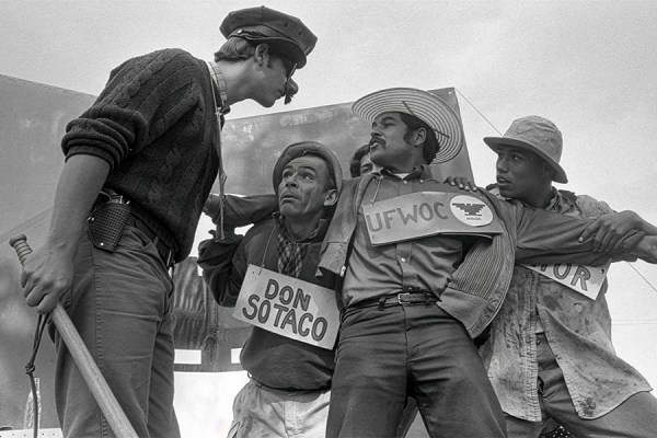 Black and white photo of an El Teatro Campesino outdoor theatrical performance supporting farmworker rights. Three actors hold signs in support of the farmworkers, looking defiantly at another actor dressed as a police officer.