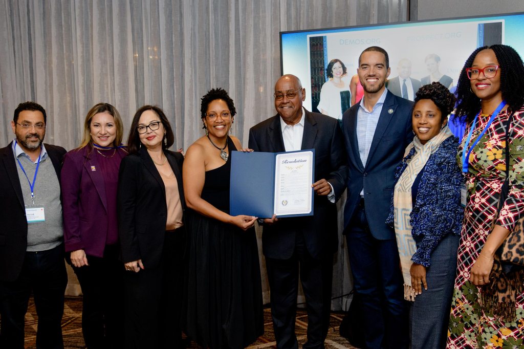 A group of professional delegates poses together in front of a Demos presentation screen as two leaders hold up an official framed resolution certificate during a democracy and data conference event.