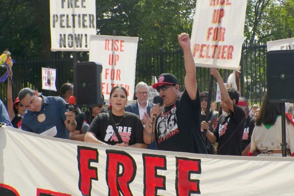 Indigenous activists holding a microphone and raising a fist at a rally with signs reading Free Peltier Now and Rise Up for Peltier.