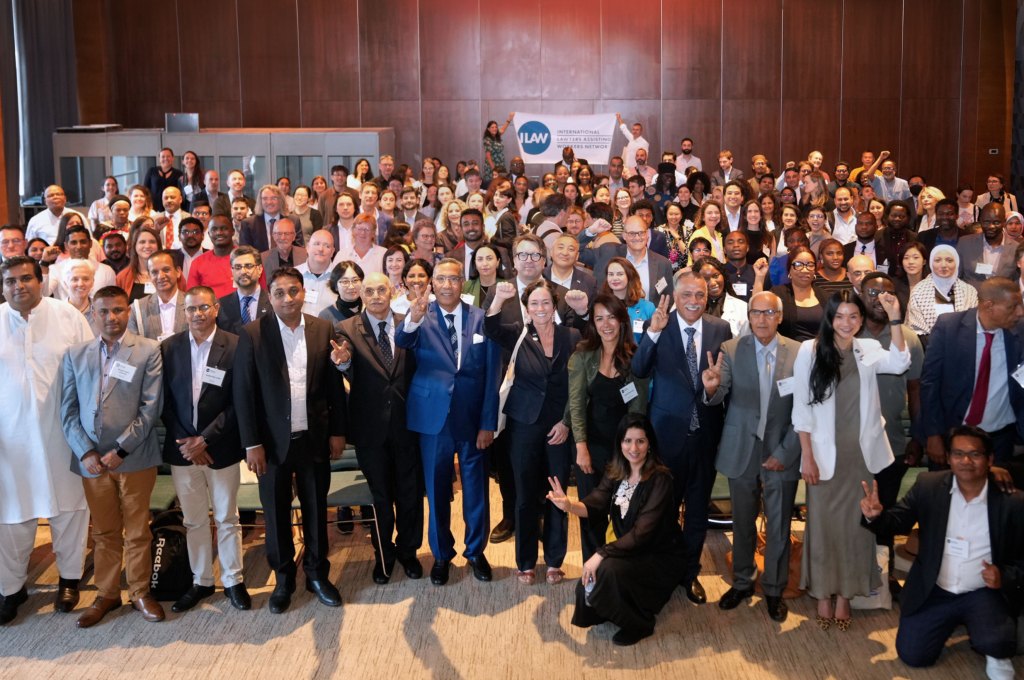 A large, diverse group of legal professionals poses for a photo in a wood-paneled conference hall. In the background, attendees hold a banner for the ILAW International Lawyers Assisting Workers Network, celebrating their global advocacy for workers' rights.