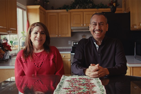 Smiling Latino woman in red sweater and laughing Latino man in black shirt sitting at a dining table in a kitchen.