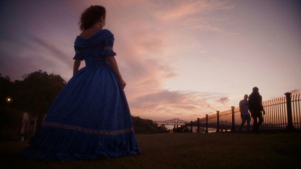 Woman in a blue Southern belle gown looking toward a bridge during a purple and orange sunset. Two people on the bridge look back at her.