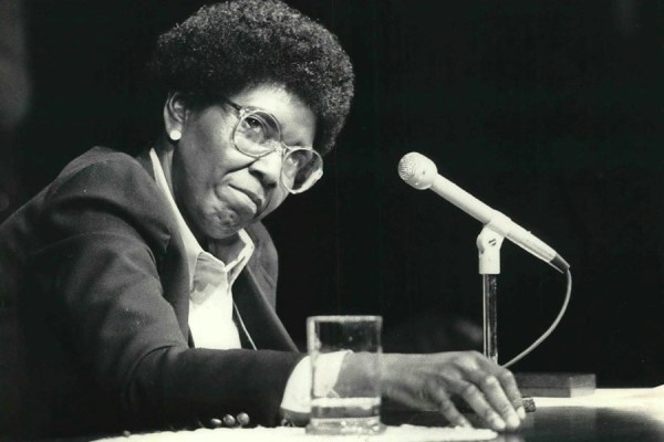 Black-and-white portrait of Congresswoman Barbara Jordan with glasses and afro hairstyle seated at a table with a microphone