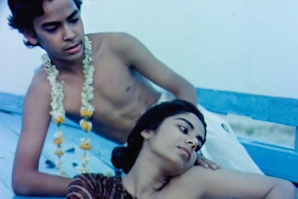 Shirtless young man wearing a flower garland leaning near a young woman resting her arm on a blue wooden surface.