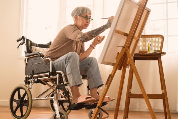 An Asian woman with short grey hair and tattooed arms sits in a wheelchair while painting on a canvas supported by a wooden easel in a brightly lit studio space, illustrating accessibility and support for disabled artists in philanthropy.