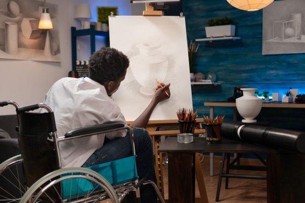 A Black male artist sitting in a wheelchair sketches a still-life vase on a canvas easel inside a home art studio. The scene illustrates themes of accessibility and supporting disabled artists through philanthropy.