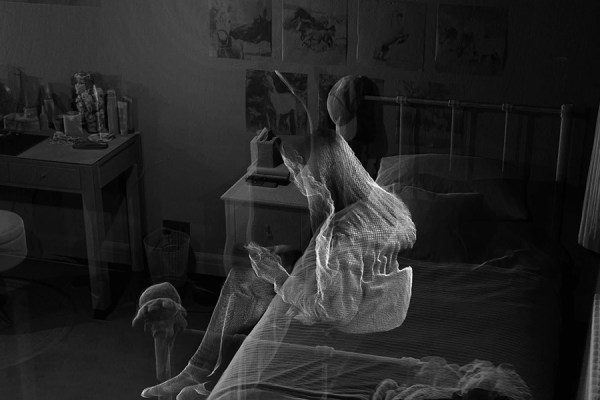 A translucent, glowing 3D wireframe figure sitting on a bed inside a dimly lit bedroom with a desk and nightstand.