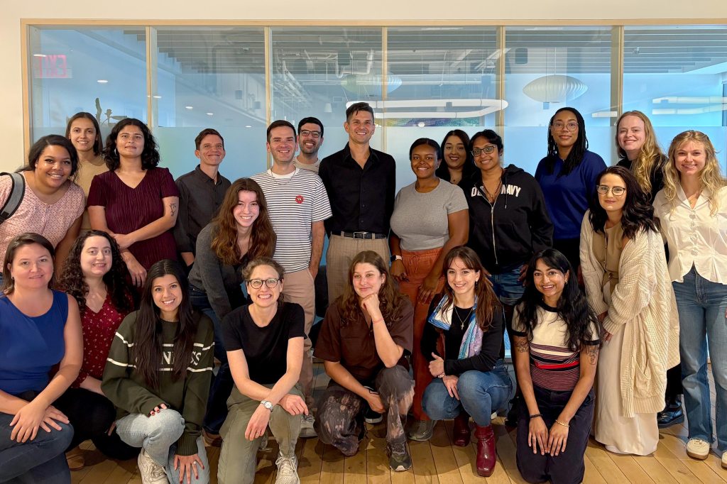 A diverse team from A Better Balance poses together inside a modern office space with large glass windows. The group, advocating for workplace equality and caregiving rights to support democracy, stands and kneels in multiple rows while smiling at the camera.