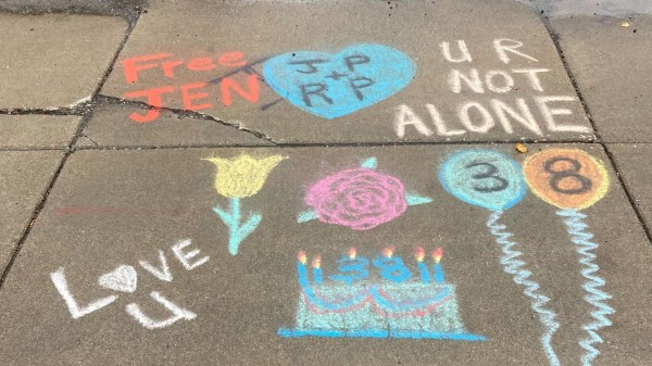 Sidewalk chalk art displaying supportive messages like "Free Jen" and "U R NOT ALONE," alongside birthday drawings including flowers, balloons with "38," and a cake, symbolizing community advocacy and equal access to justice.