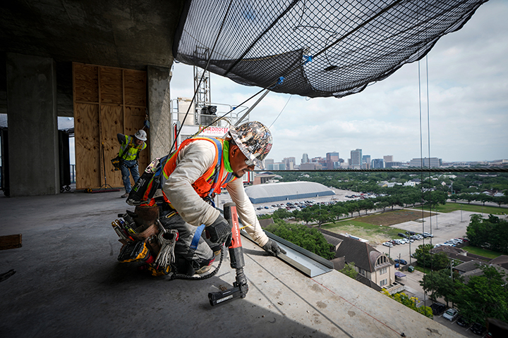 A construction worker wearing a hard hat, safety harness, and tool belt uses power tools on an upper floor of a high-rise building under construction. The cityscape is visible in the background, representing skilled immigrant labor powering infrastructure development.