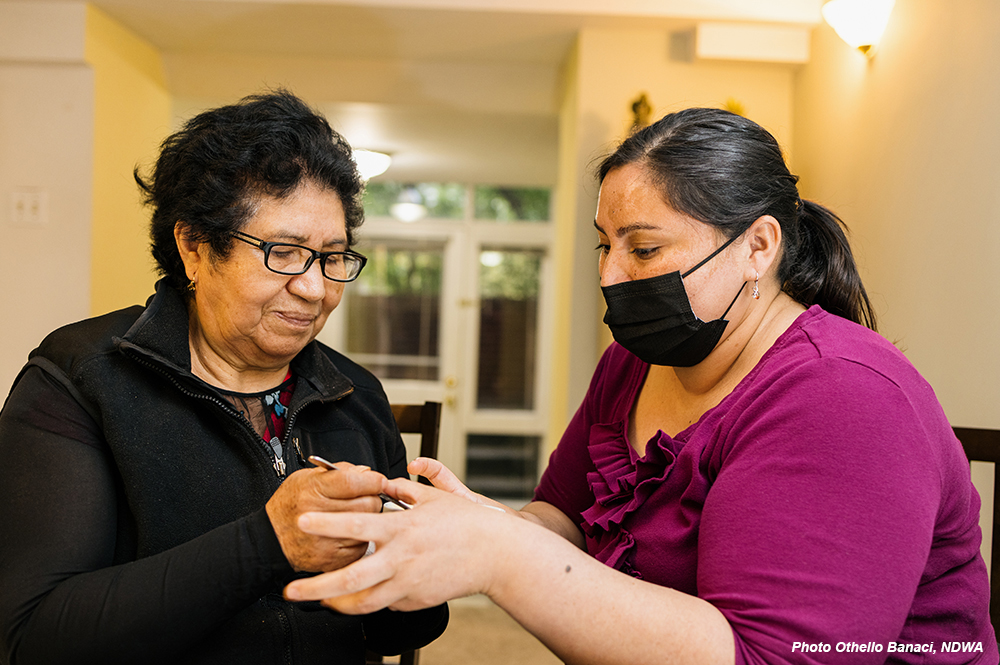 An elderly woman with glasses carefully performs a manicure on a younger woman wearing a black face mask inside a home setting, illustrating essential caregiving, personal service labor, and immigrant contributions to the workforce.