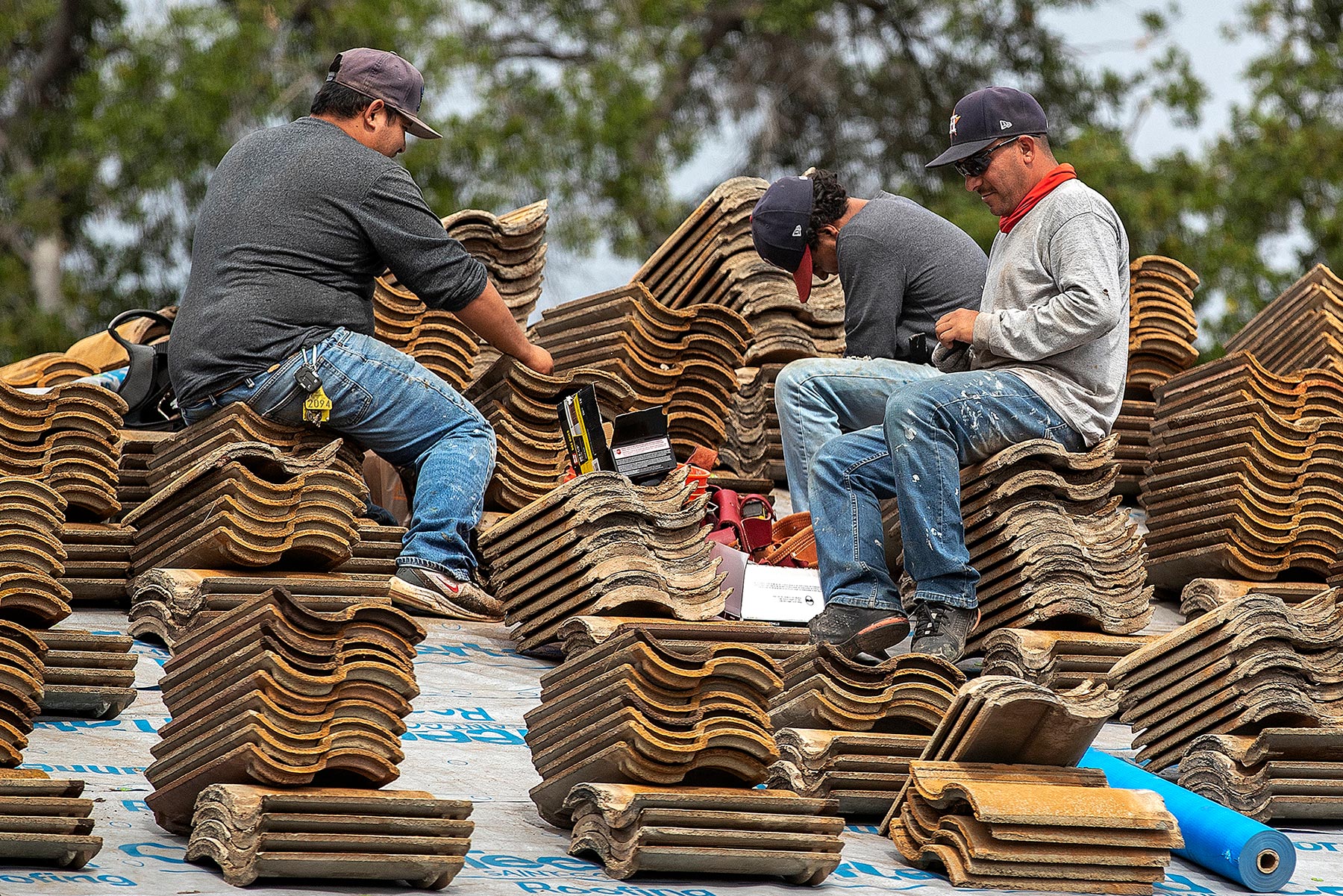 Three construction workers sit among stacked clay roof tiles while installing or repairing a rooftop. The image highlights skilled manual labor and immigrant contributions to the building trades and workforce.