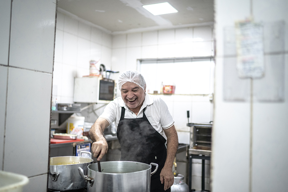 A cheerful Hispanic chef wearing a hairnet and apron laughs while stirring a large steaming pot of food in a commercial kitchen, highlighting immigrant talent powering the culinary and food service industry.