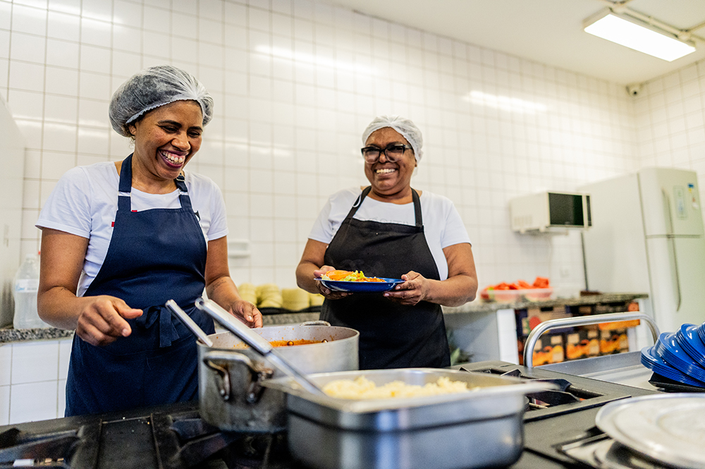 Two smiling female kitchen workers wearing hairnets and aprons prepare and plate meals in a tiled commercial kitchen, highlighting the essential contributions of immigrant talent to the food service and hospitality workforce.