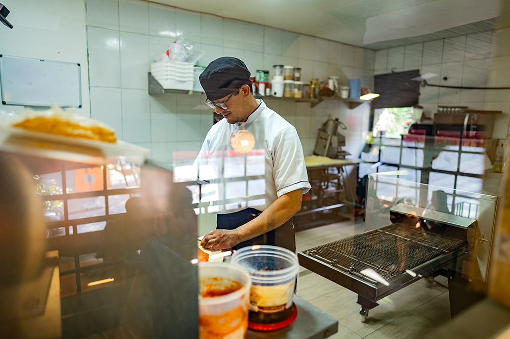 A chef wearing glasses, a white uniform, and a black cap prepares food behind a glass display counter in a clean commercial kitchen, showcasing skilled immigrant labor and culinary expertise in the food service industry.