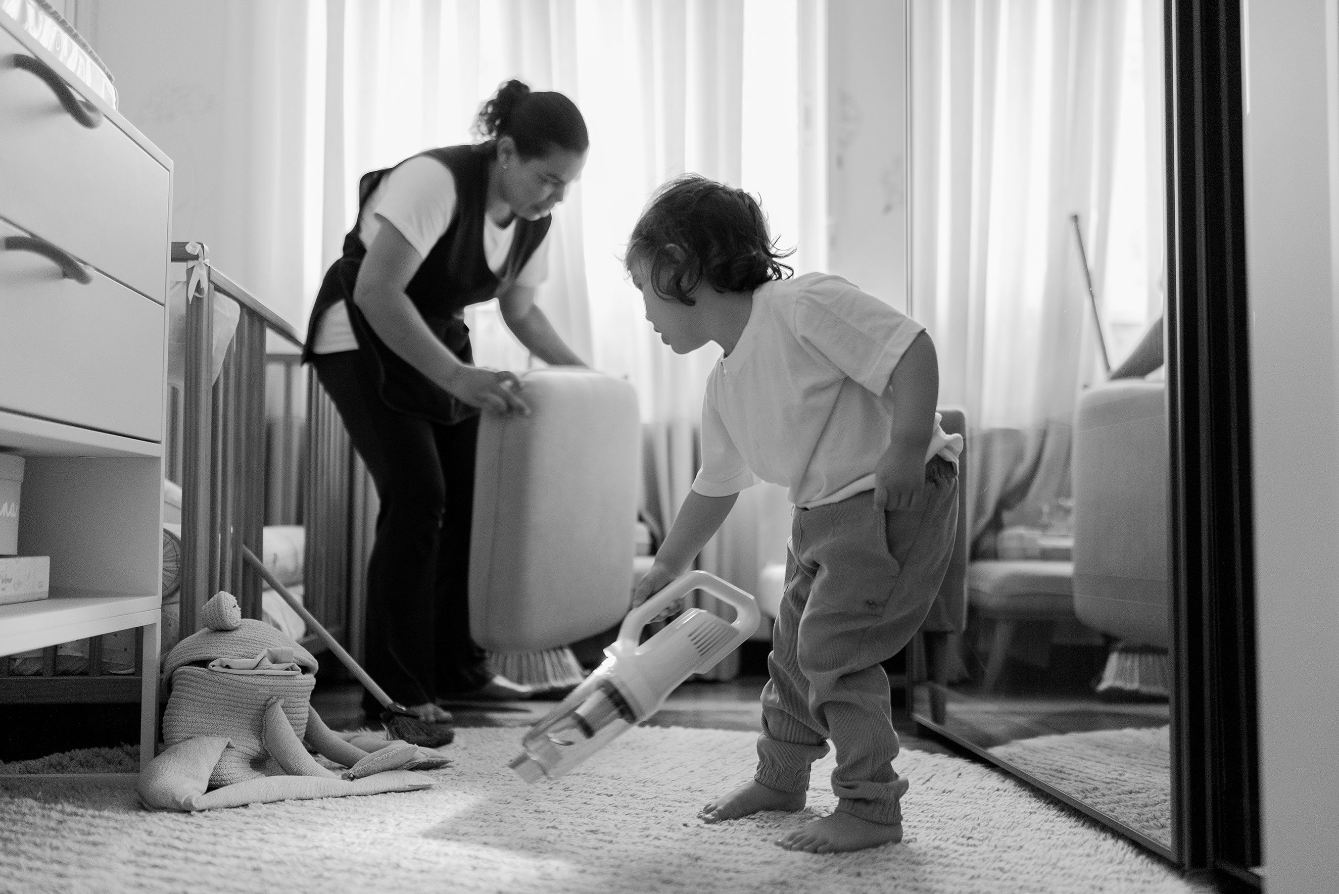 A black-and-white photo of a domestic worker organizing a bedroom while a young child playfully holds a small handheld vacuum cleaner, highlighting essential childcare, domestic labor, and immigrant talent in home environments.