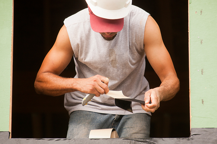 A construction worker wearing a white hard hat cuts and prepares weatherproofing tape with a utility knife while framing a window opening, highlighting skilled trades and immigrant labor driving building development.