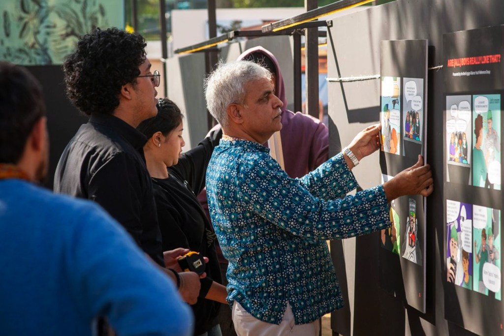 An older man in a blue patterned shirt sets up an educational comic exhibition display with young advocates nearby. The MenEngage Alliance initiative focuses on engaging men and boys to promote gender equality and work toward a just society.