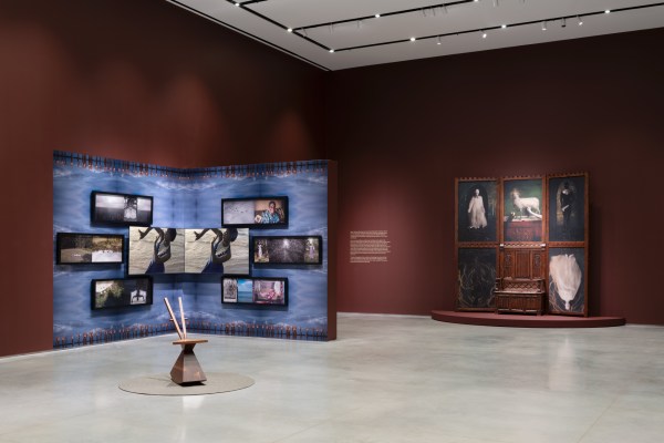 A museum gallery displaying two large installations. On the left, multiple screens show various images on a blue patterned wall, with a light wooden chair sculpture in the foreground. On the right, a dark wooden gothic pew connected to several framed paintings stands on a circular platform.