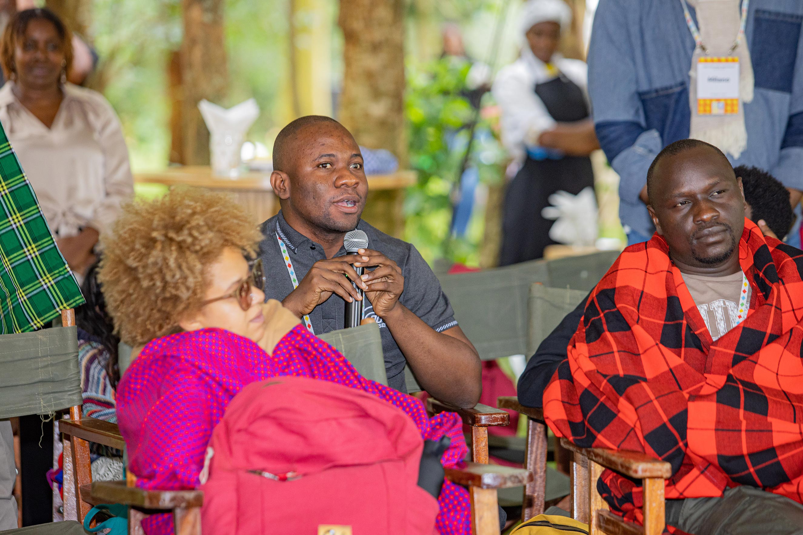 Rasak Adekoya speaking into a microphone while seated among participants wearing traditional blankets during an outdoor workshop focused on disability inclusion.