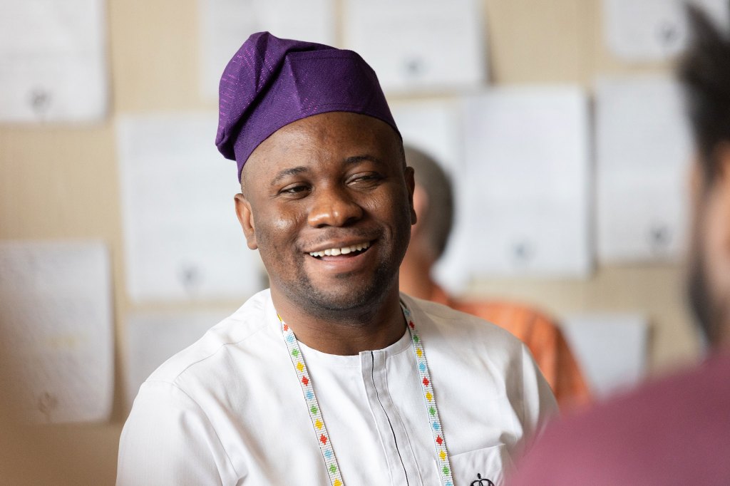 Rasak Adekoya smiling while wearing traditional Nigerian attire and a purple filas cap, engaging in a discussion about redesigning systems for disability inclusion.