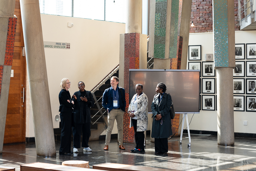 A group of professionals standing and conversing in a modern, brightly lit lobby with mosaic-tiled pillars and framed portrait photos, taken during a visit to South Africa.