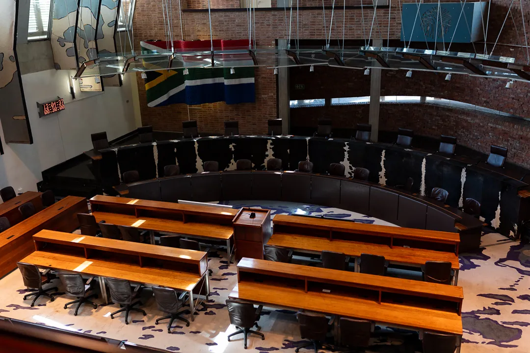 An overhead view of the empty Constitutional Courtroom of South Africa, featuring wooden desks, a curved bench with cowhide panels, brick walls, and the South African flag hanging in the background.