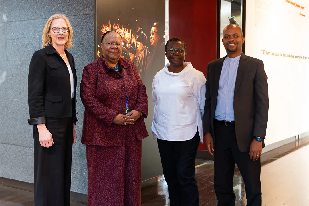 Four delegates posing for a photo inside a modern exhibit hall during a visit to South Africa, standing in front of wall displays and historical photos.