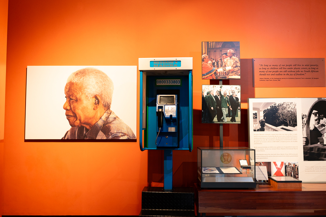 A museum exhibit dedicated to Nelson Mandela, featuring a blue "Message 4 Madiba" payphone booth against an orange wall, flanked by portrait photos, quotes, and historical display cases from South Africa.