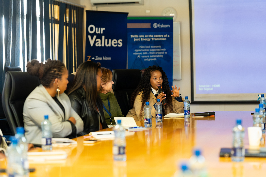 A woman speaking into a microphone while seated at a long conference table with colleagues during a meeting on South Africa's Just Energy Transition, in front of an Eskom banner.