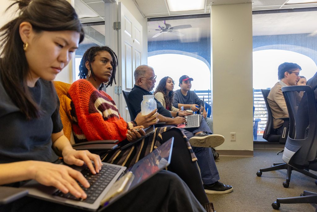 Diverse team of journalists and staff working on laptops in a Deep South Today newsroom meeting, focused on local community reporting.