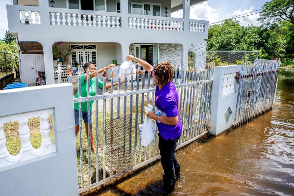 A volunteer standing in flooded street waters hands bags of essential aid and supplies over a residential fence to a resident following a natural disaster relief effort.