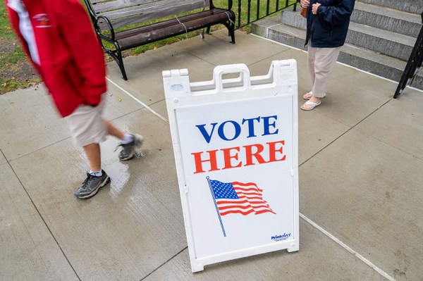 A white A-frame sign with an American flag and bold text reading "VOTE HERE" stands on a concrete sidewalk outside a polling location as voters walk past during an election.