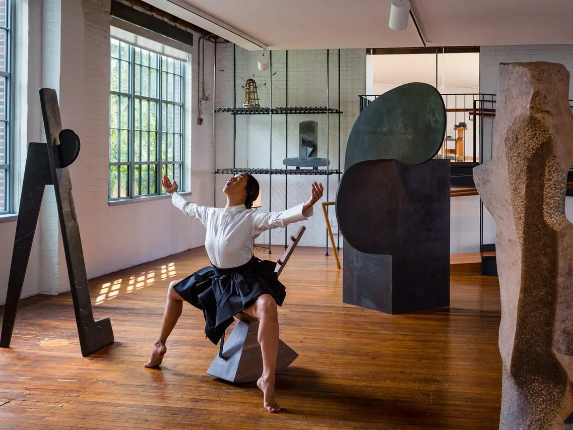 A performance of a dancer sitting on a sculptural rocking chair, with her feet pointed and arms spread outward. She poses amidst large ceramic sculptures in a brightly-lit gallery with open windows.
