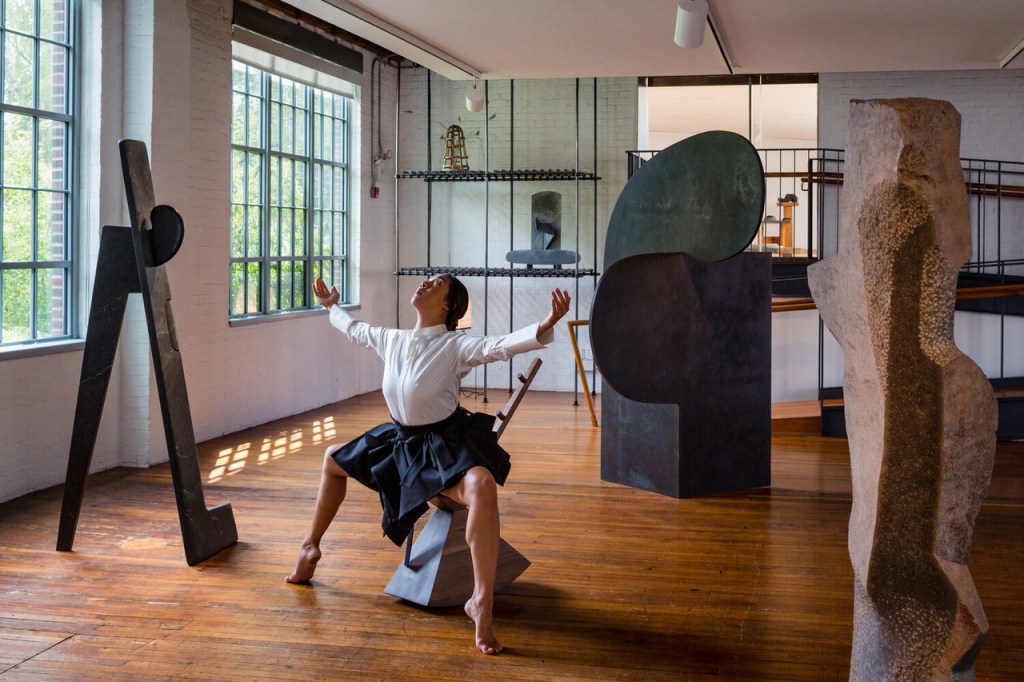 A performance of a dancer sitting on a sculptural rocking chair, with her feet pointed and arms spread outward. She poses amidst large ceramic sculptures in a brightly-lit gallery with open windows.