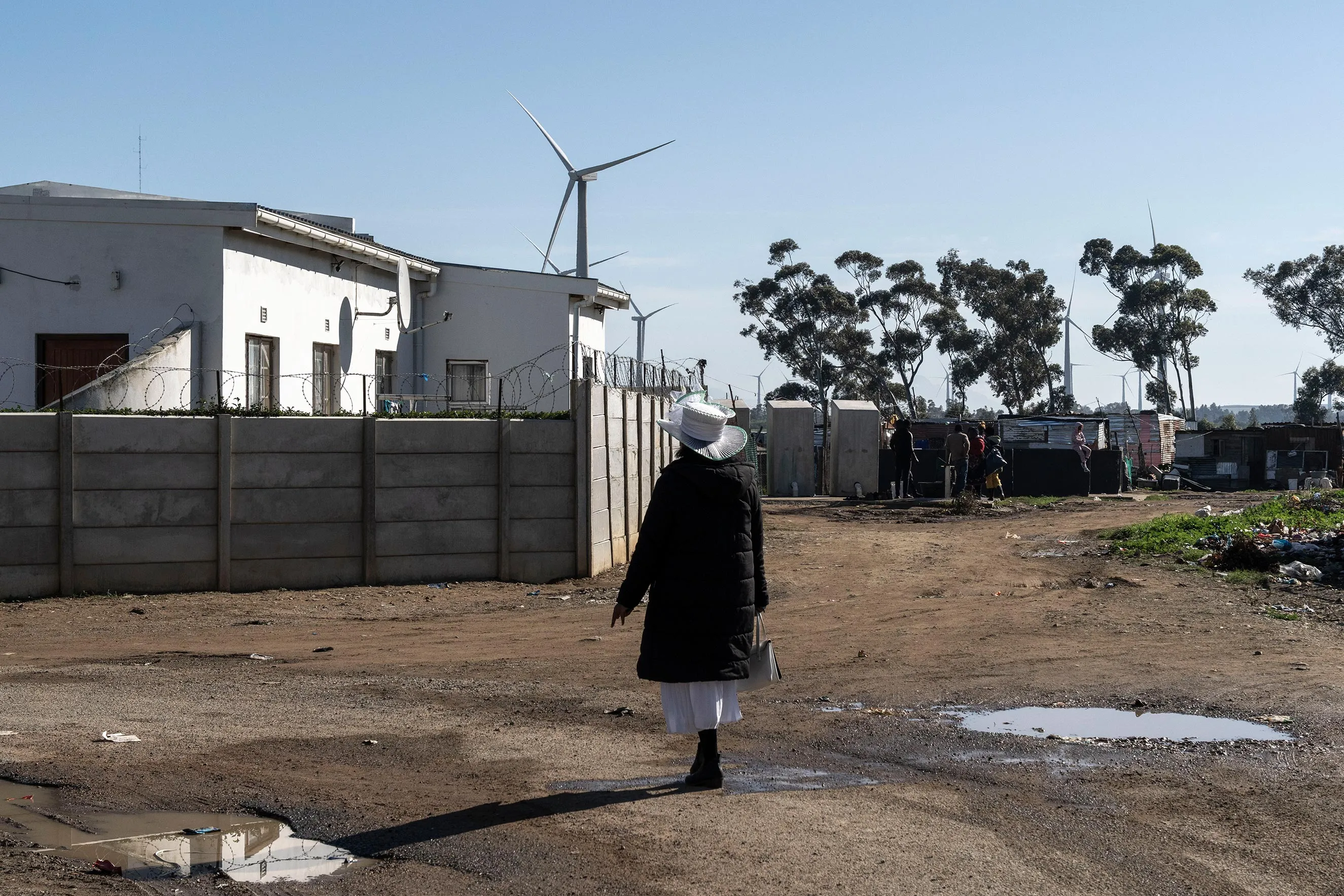 A woman in a dark coat and white hat walking down a dirt road in a South African community[cite: 21], with a walled building, informal dwellings, and tall wind turbines visible in the background under a clear sky.