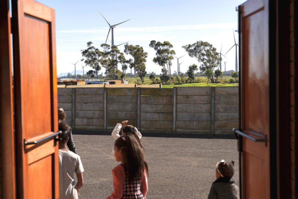 View through open wooden doors of children standing in a courtyard facing a concrete wall, with tall wind turbines and trees visible in the distance under a clear sky, representing South Africa’s community energy transition.