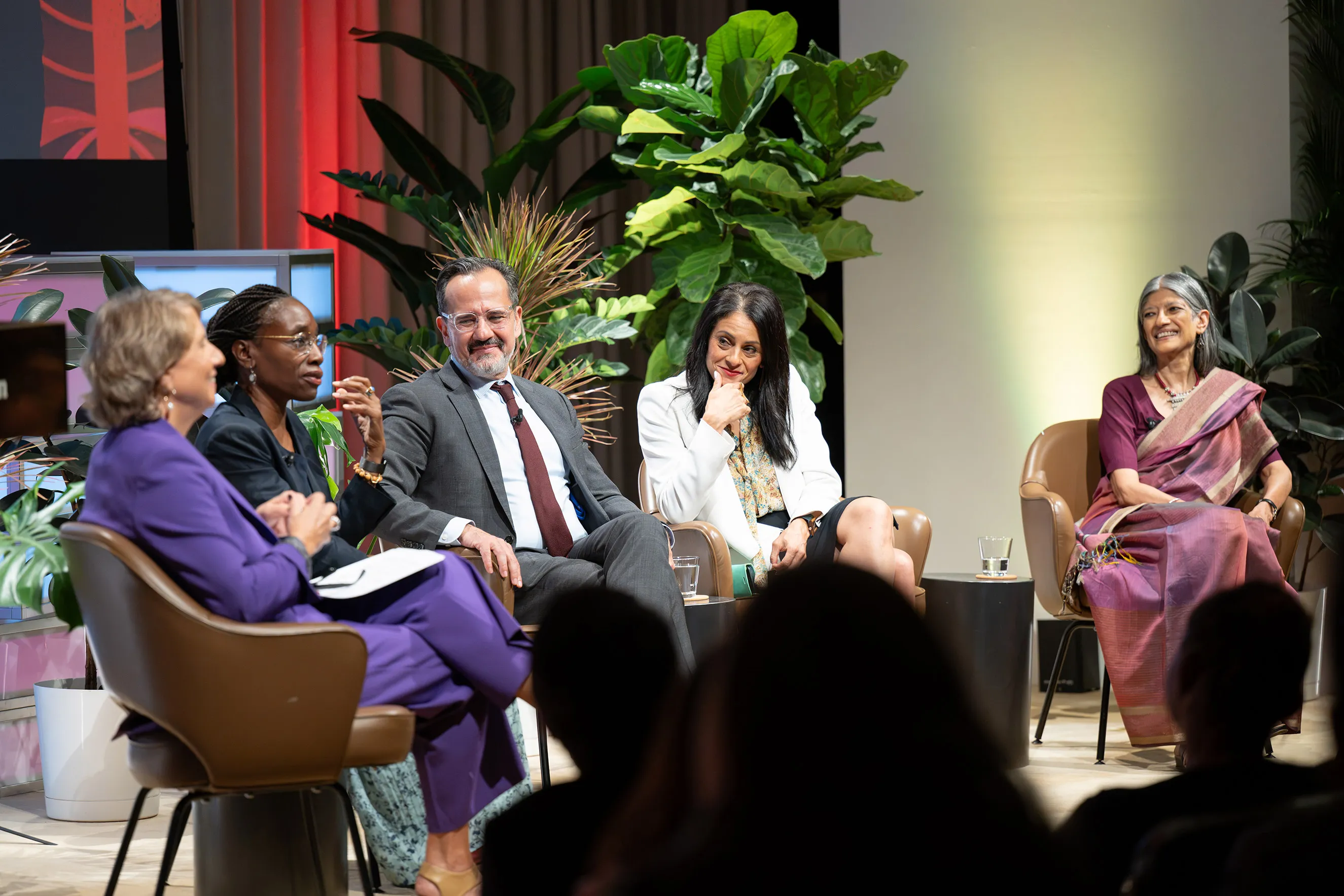 Five panel speakers seated on stage in armchairs surrounded by lush green potted plants during an event, engaged in conversation in front of an audience.