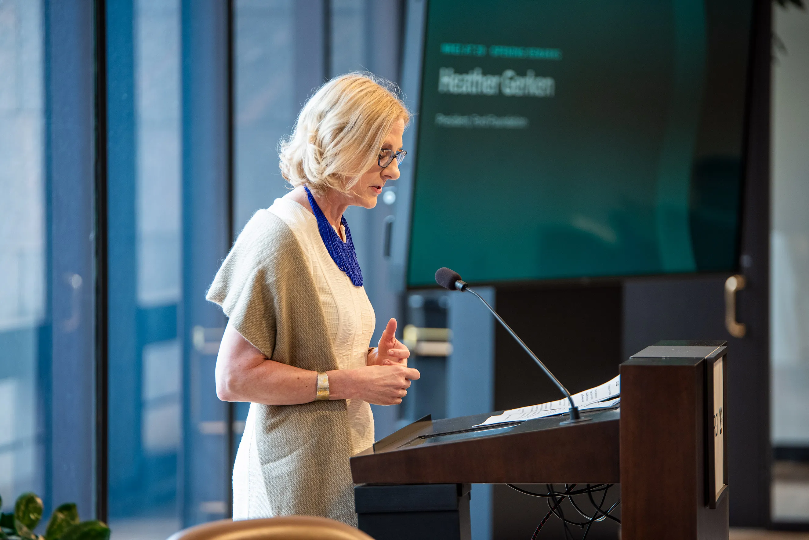 Heather Gerken speaking at a podium during an event, with a screen displaying her name and title in the background.