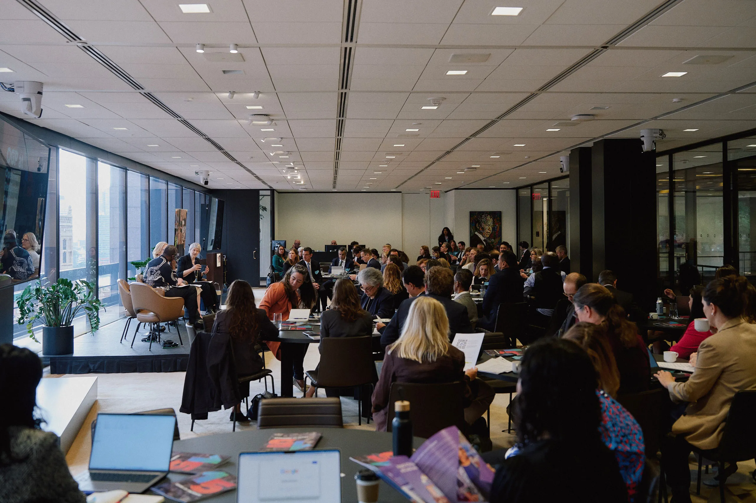 Wide shot of an audience seated at round tables facing a stage panel presentation in a modern conference room with large windows overlooking city views.
