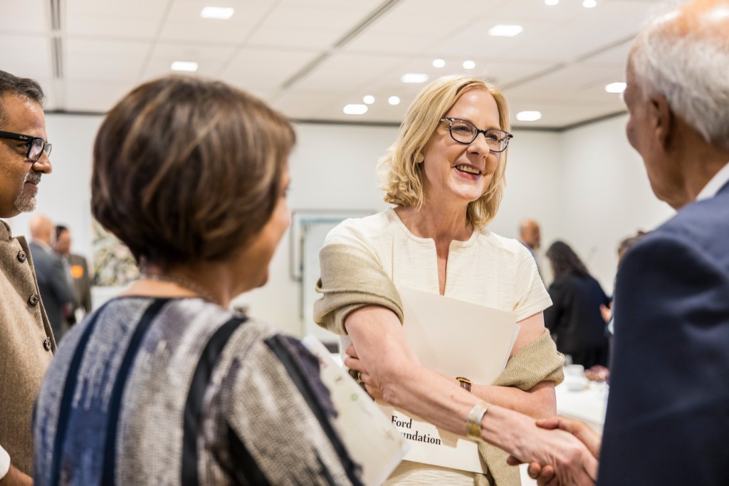 Heather Gerken smiling and shaking hands with an attendee while holding a Ford Foundation folder during a networking gathering.
